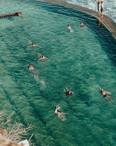 High angle of unrecognizable tourists in swimsuit swimming in pool near pier with railing and waving ocean with rocky formations in sunny summer day